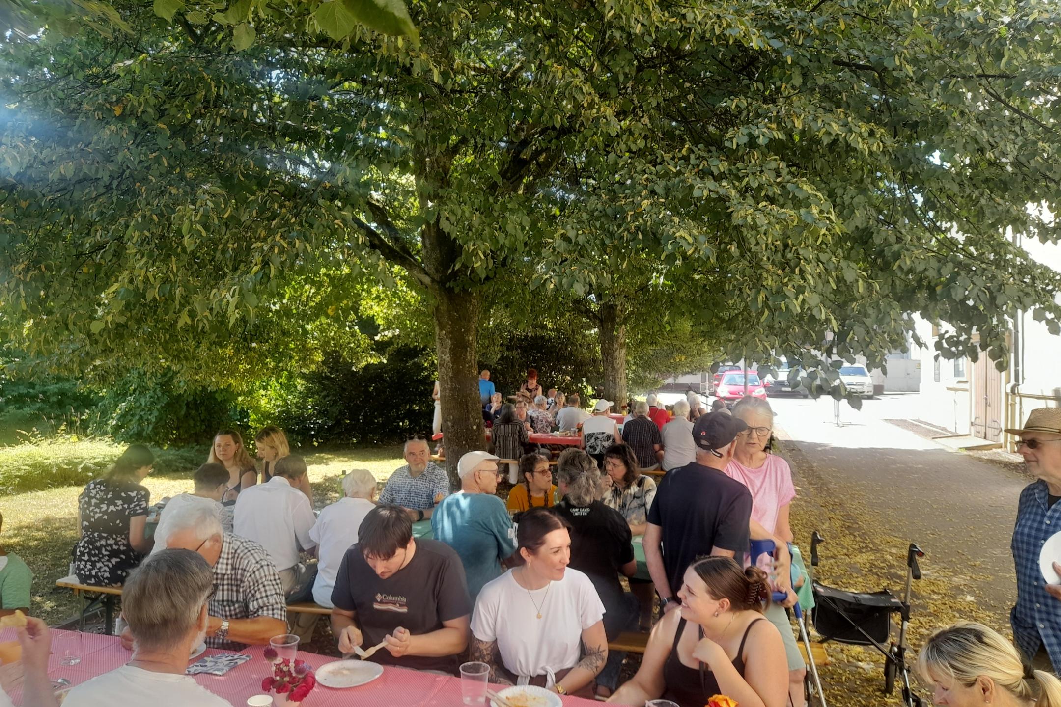Unter den Bäumen sitzen Menschen  an Tischen, essen und unterhalten sich