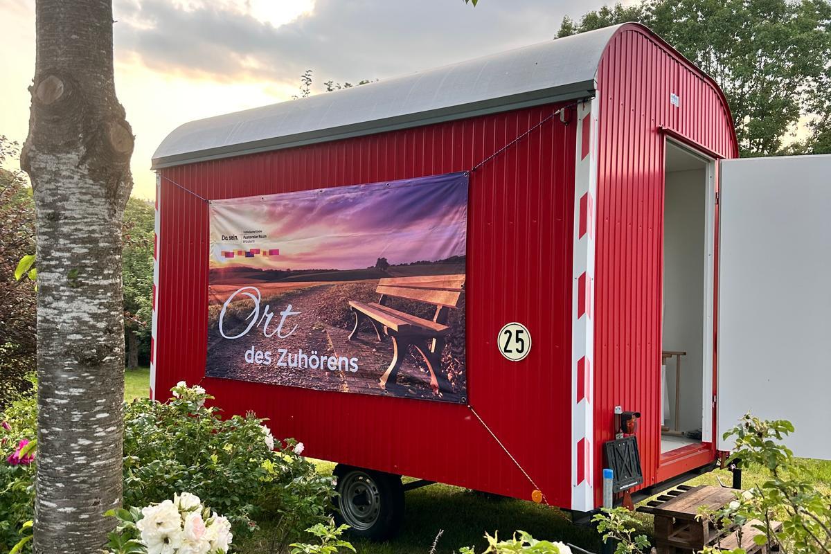 Ein Roter Bauwagen mit der Aufschrift 'Ort des Zuhörens' inmitten von blühenden Büschen und einem Baum