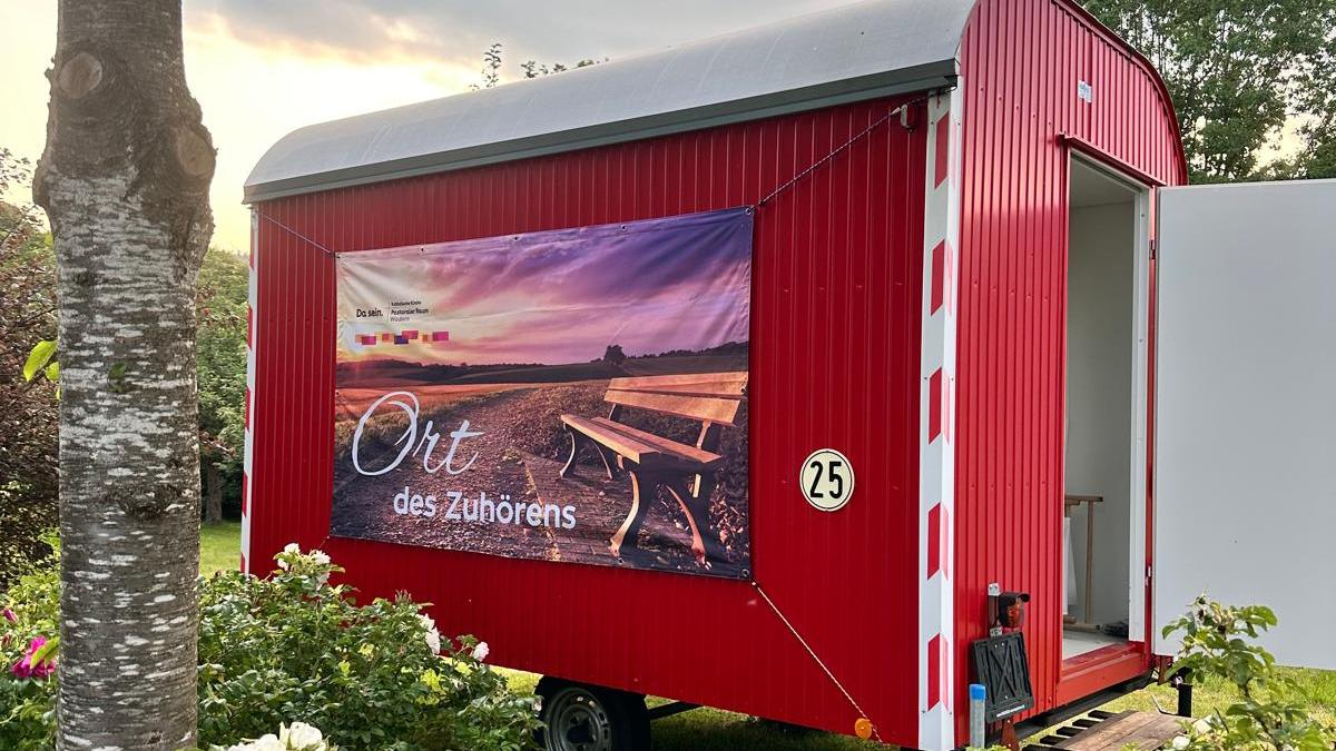 Ein Roter Bauwagen mit der Aufschrift 'Ort des Zuhörens' inmitten von blühenden Büschen und einem Baum
