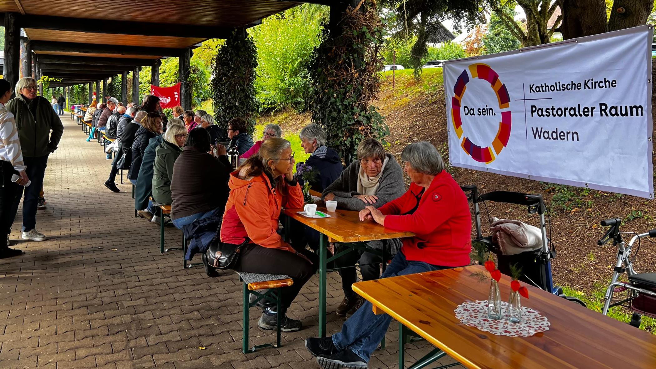 Menschen sitzen gemeinsam an einer Tafel von Biertischen davor das Plakat vom Pastoralen Raum Wadern