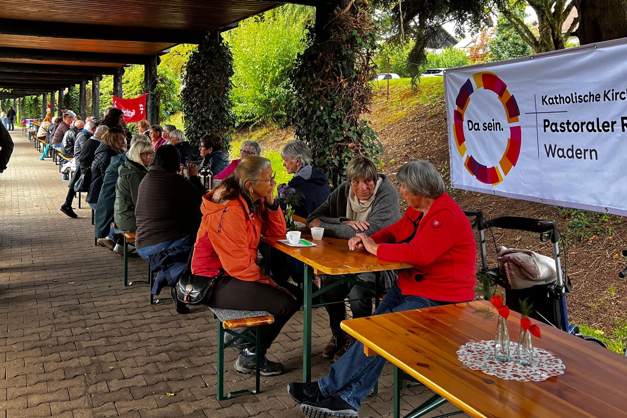Menschen sitzen gemeinsam an einer Tafel von Biertischen davor das Plakat vom Pastoralen Raum Wadern